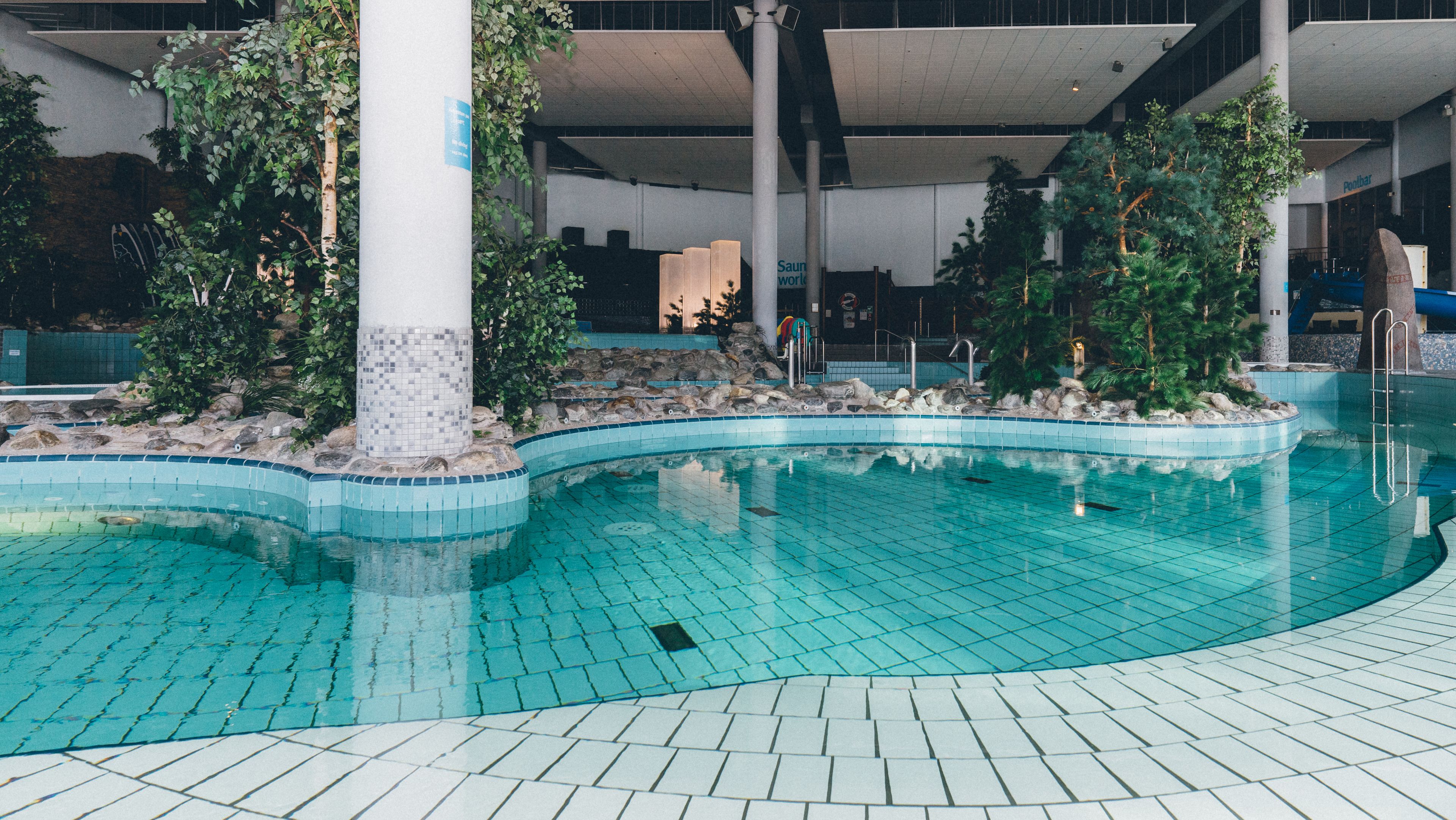 An indoor swimming pool with tiled flooring and a curved design, surrounded by rocks and greenery.