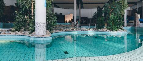 An indoor swimming pool with tiled flooring and a curved design, surrounded by rocks and greenery.