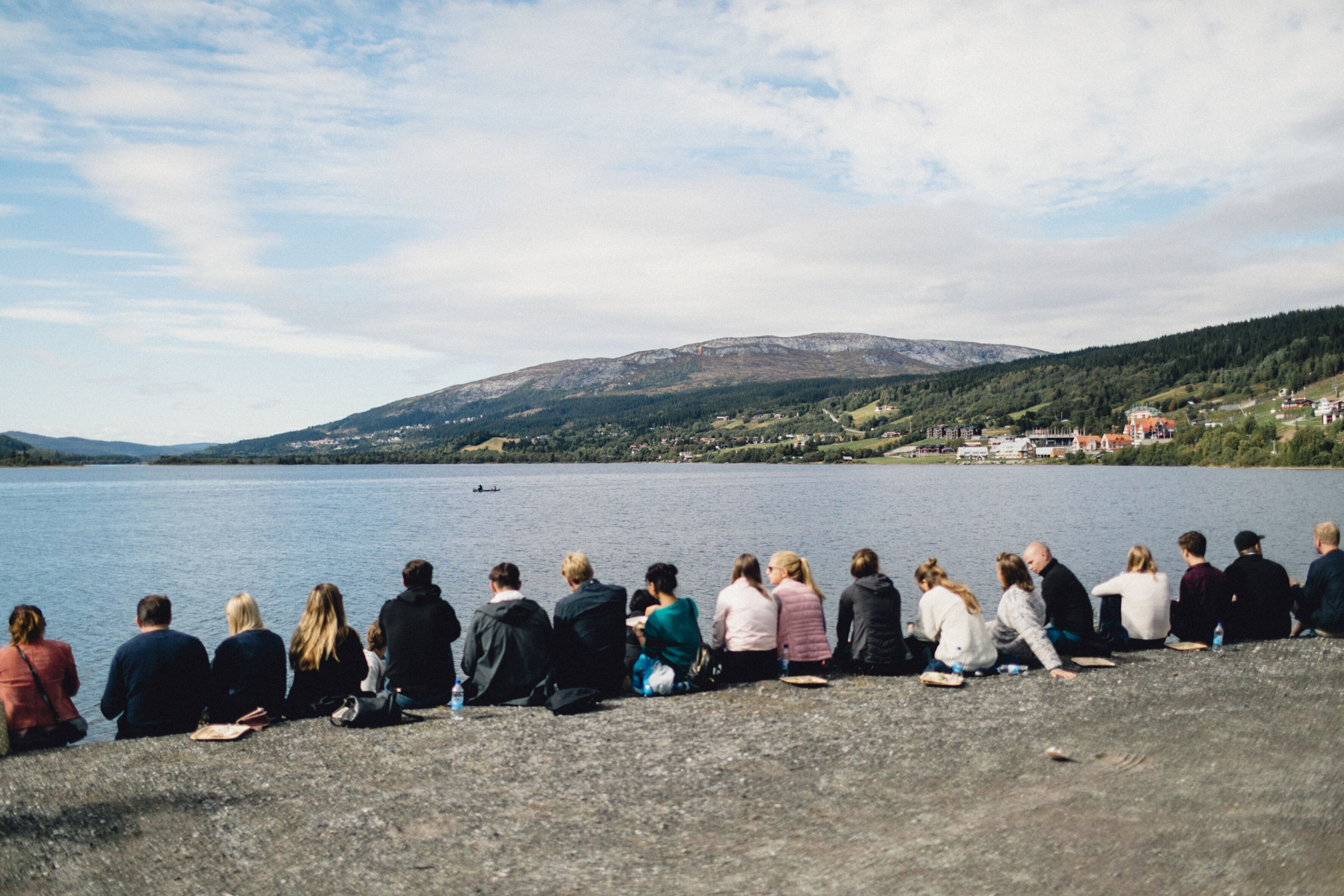 A group of people sitting on a rocky shore by a lake with a mountain in the background.