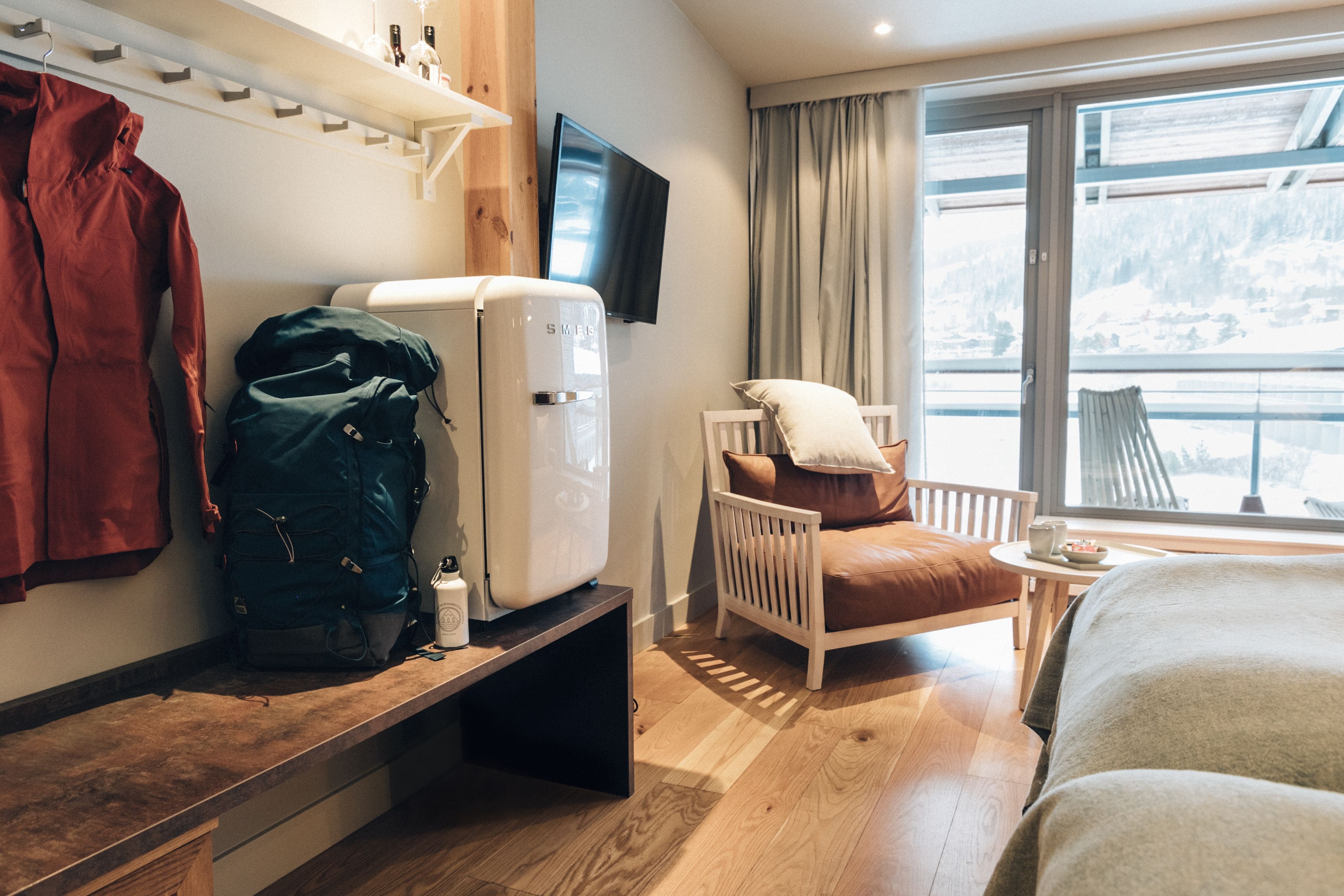 Habitación de hotel con cama, una silla con cojín, una mesita con una taza, televisor montado en la pared y ventana con vista a un paisaje nevado.