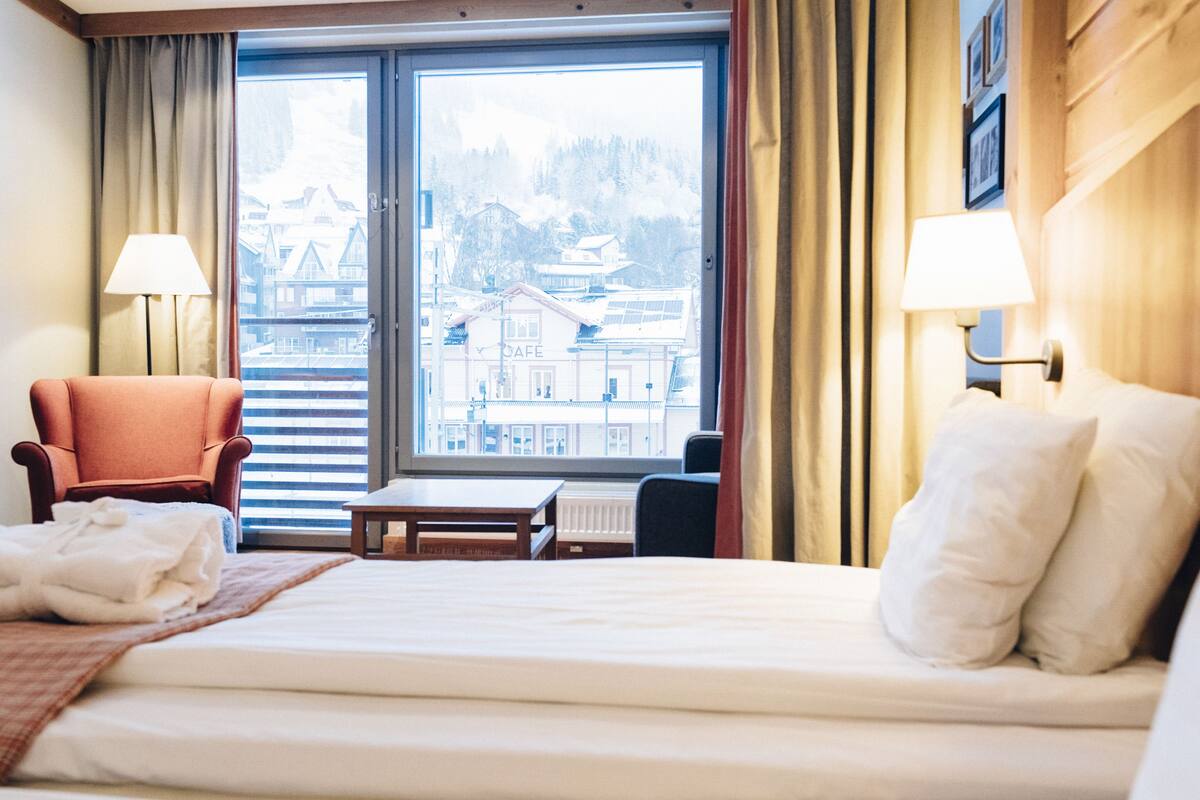 A hotel room with a bed, a chair, a table, and a view of buildings through a window.