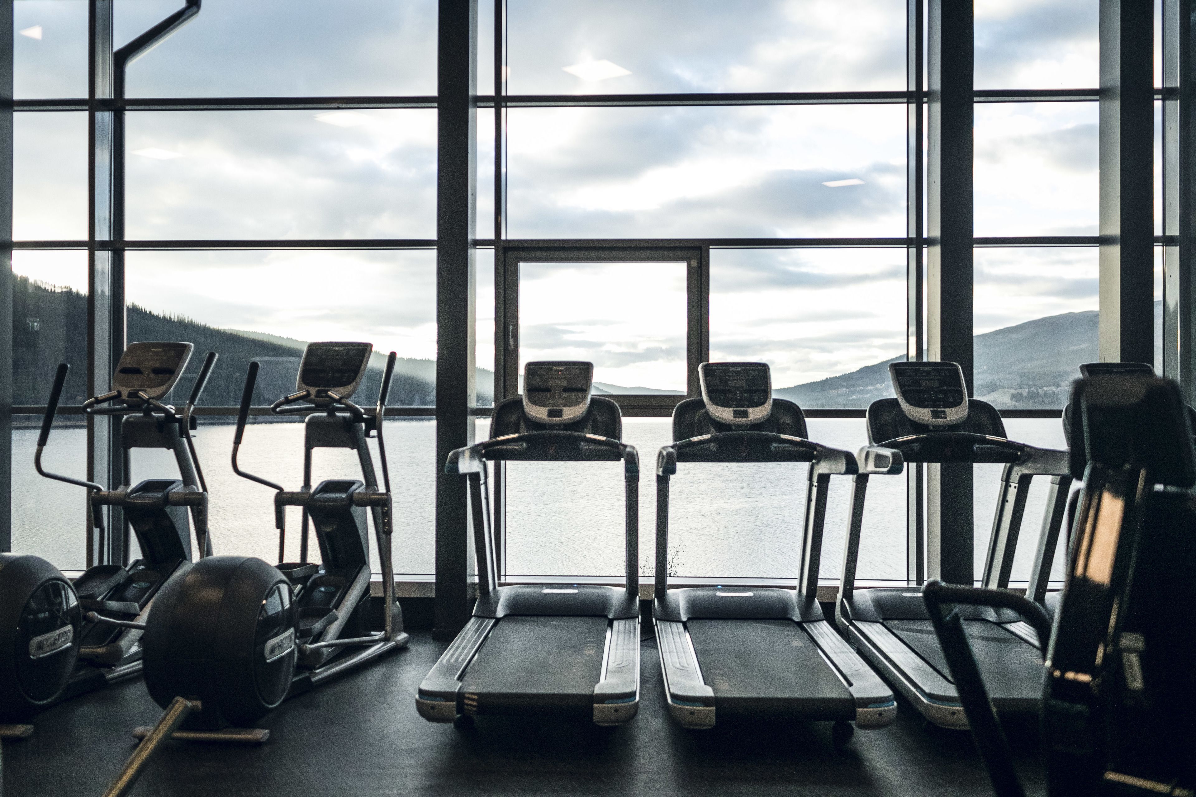 A gym with treadmills and elliptical machines, large windows, and a view of a lake and mountains.