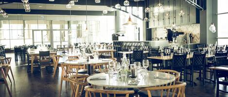 A spacious restaurant with multiple tables and chairs, large windows, and hanging spherical light fixtures.