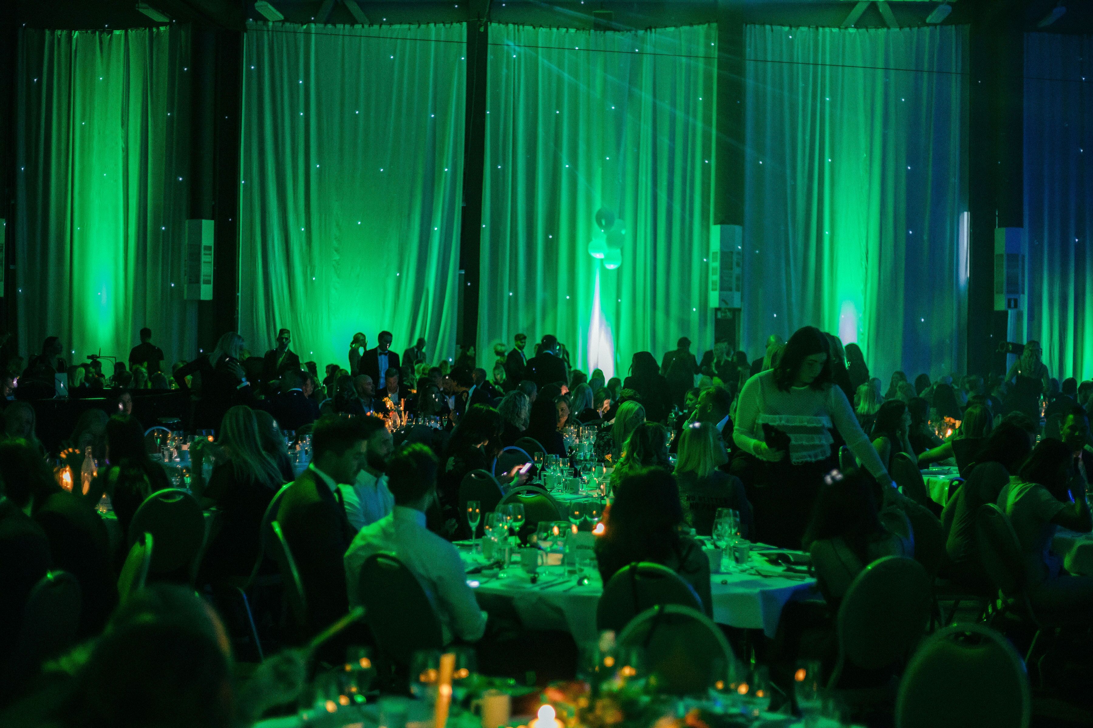 Een balzaal met gedekte tafels voor een formeel evenement, met draperende gordijnen en een menigte aanwezigen.