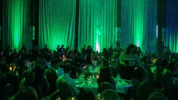 Een balzaal met gedekte tafels voor een formeel evenement, met draperende gordijnen en een menigte aanwezigen.