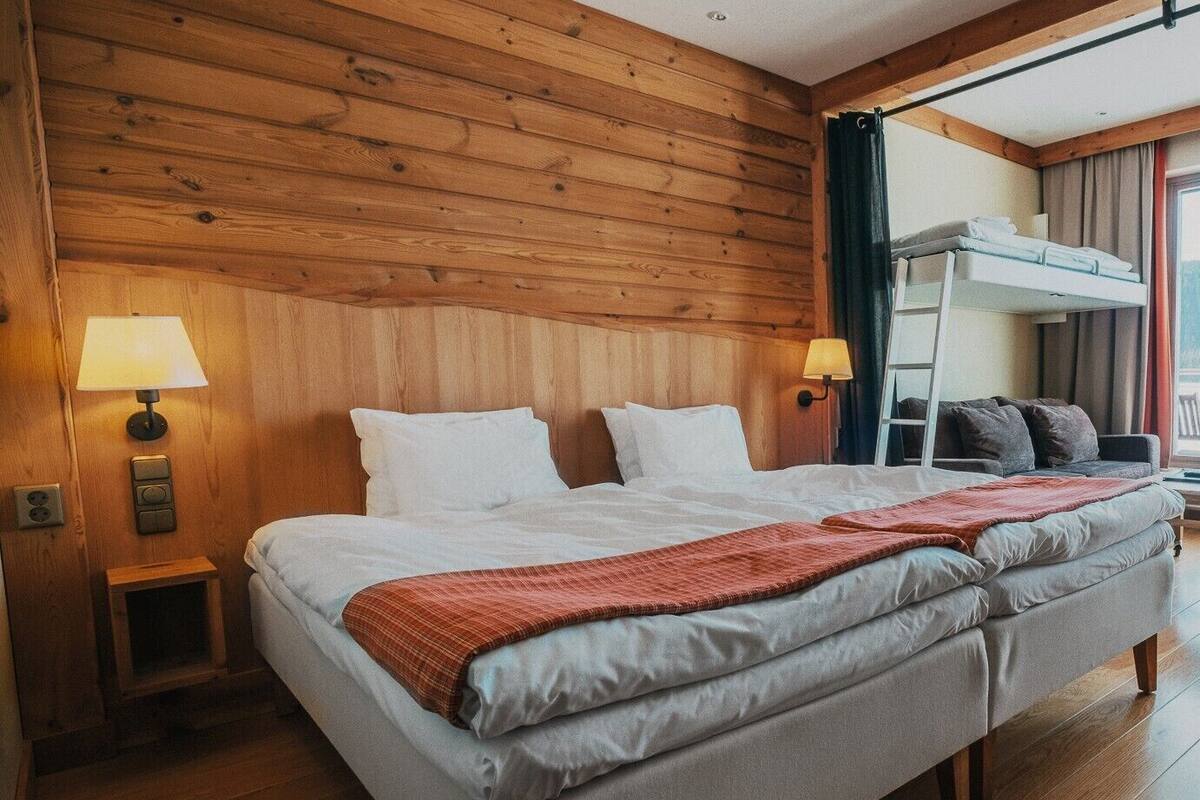 A hotel room with a wooden paneled wall, a large bed with white linens, two bedside lamps, a bunk bed, and a wooden floor.