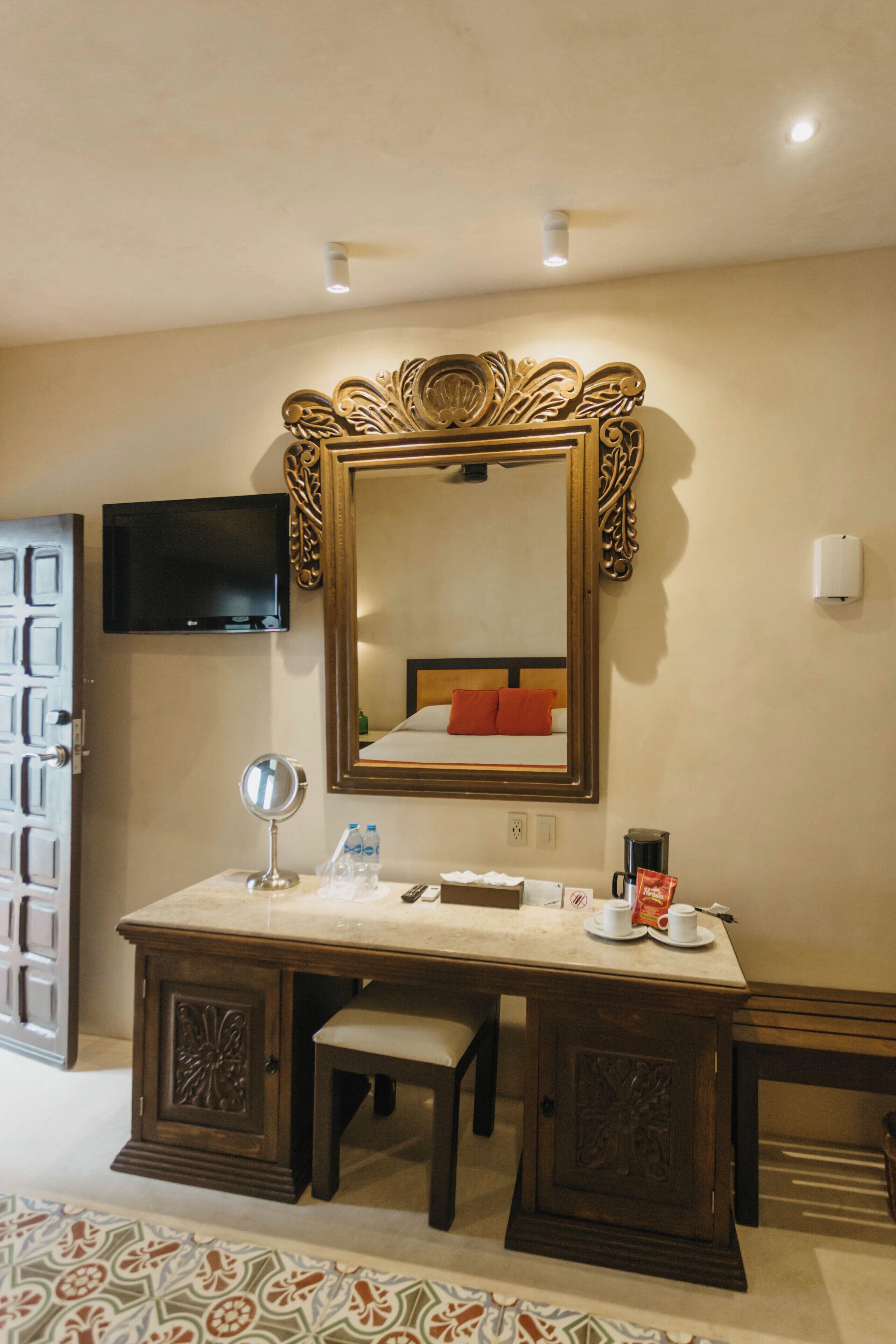 A hotel room with a large ornate mirror, a television mounted on the wall, a bedside table with a cup and a bottle, and a patterned rug on the floor.