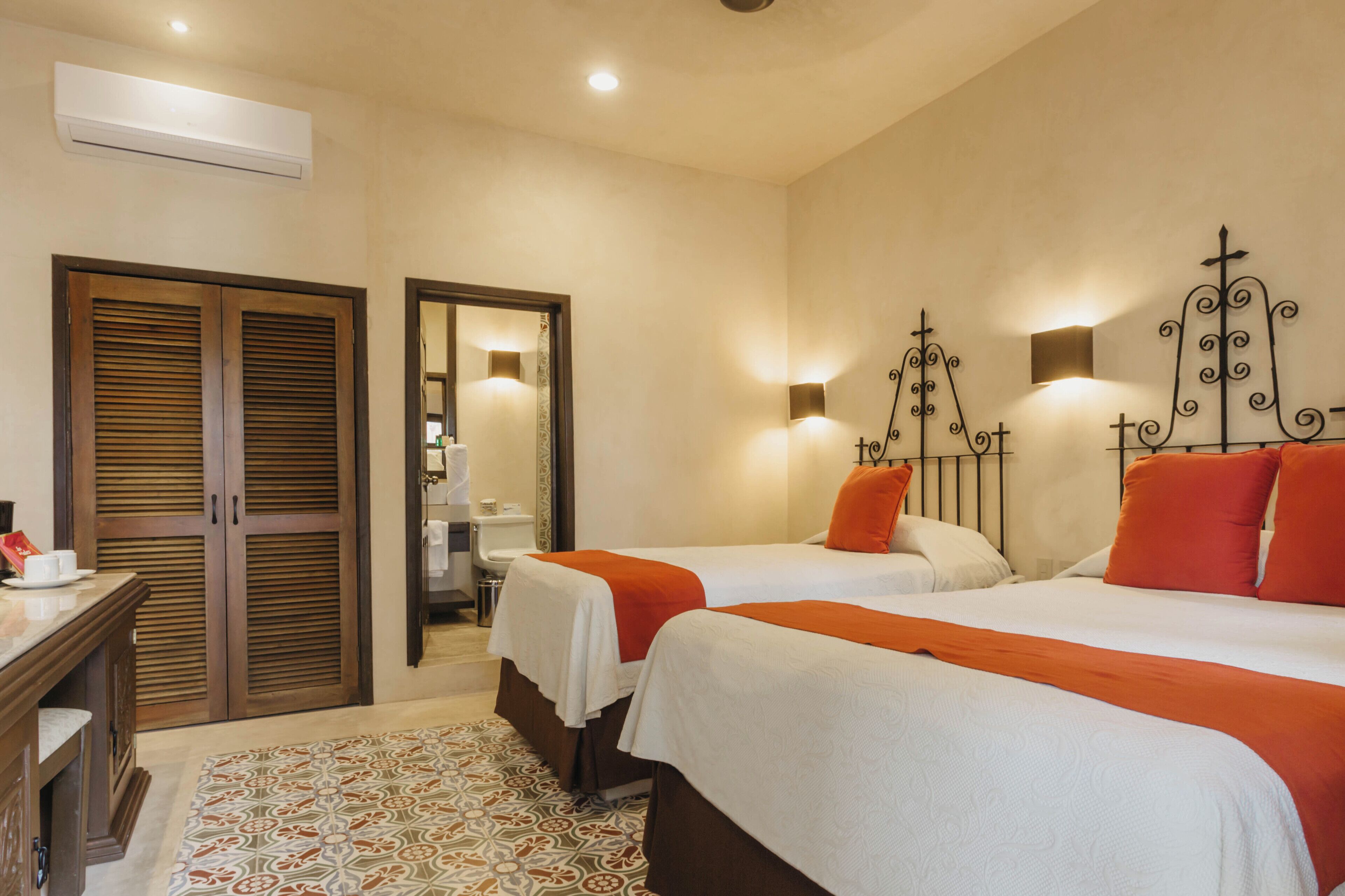 A hotel room with two beds, a wooden wardrobe, a bathroom visible through a door, and patterned floor tiles.