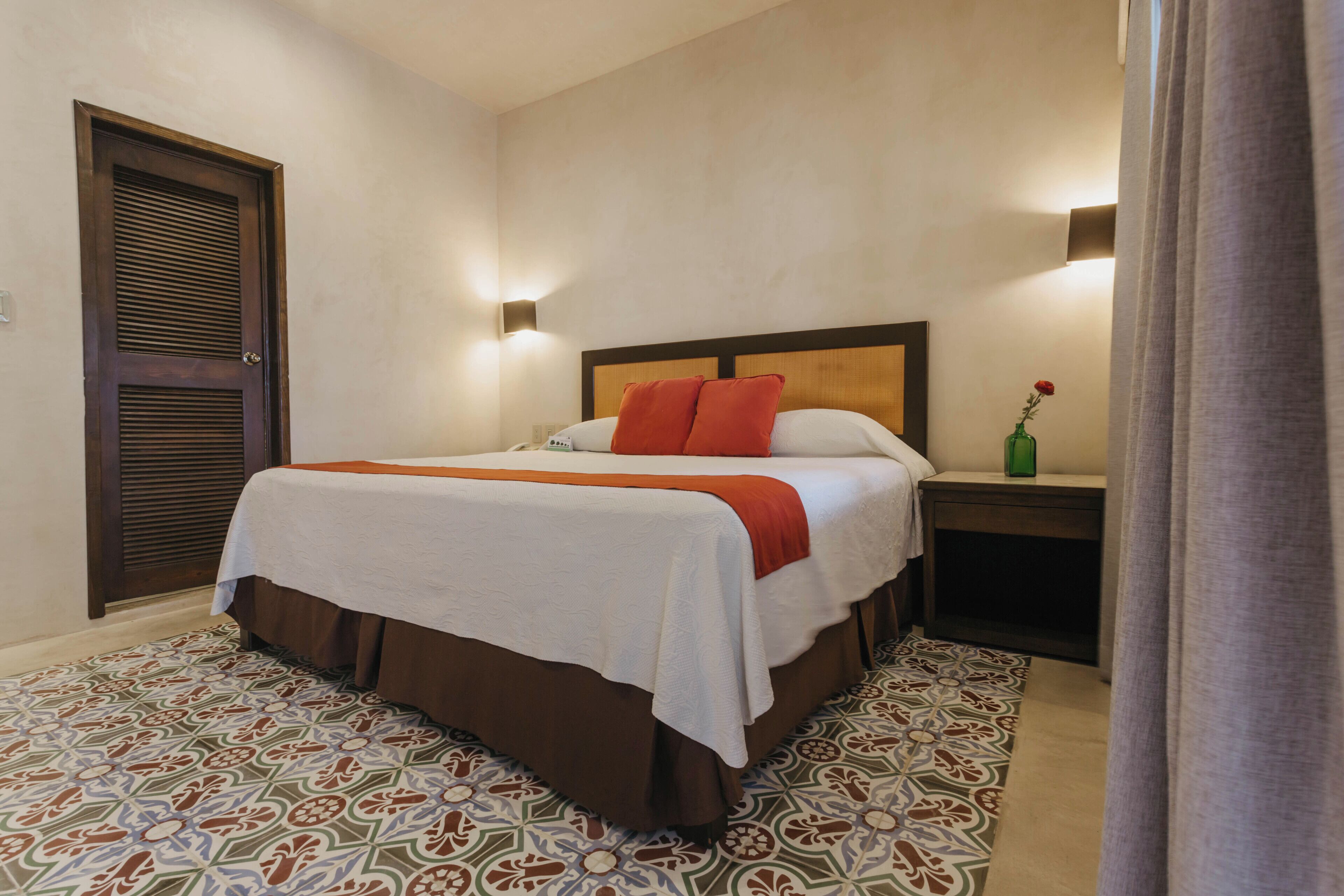 A hotel room with a bed, a nightstand, a wall-mounted lamp, and a patterned rug.