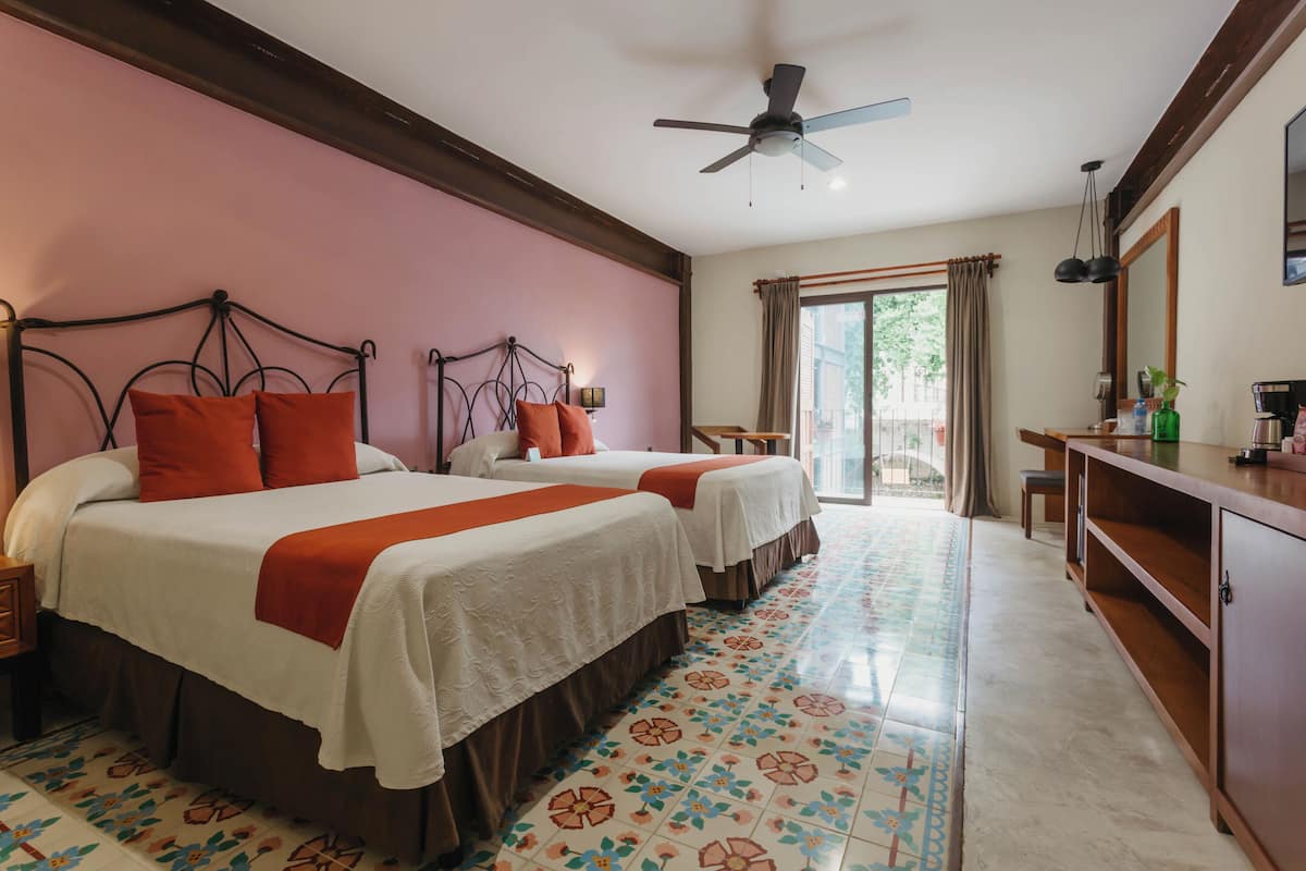 Une chambre d’hôtel avec deux lits, un ventilateur de plafond, une grande fenêtre avec des rideaux, un bureau en bois et un sol carrelé.
