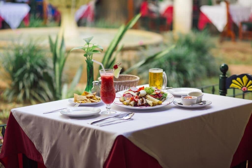 Mesa dispuesta con varios platos, una bebida y una jarra, al aire libre con plantas y banderas al fondo.