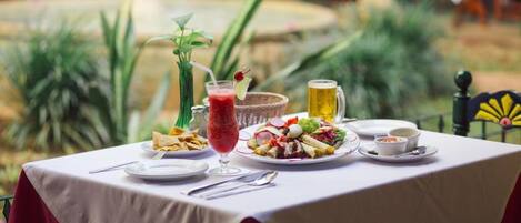 Mesa dispuesta con varios platos, una bebida y una jarra, al aire libre con plantas y banderas al fondo.