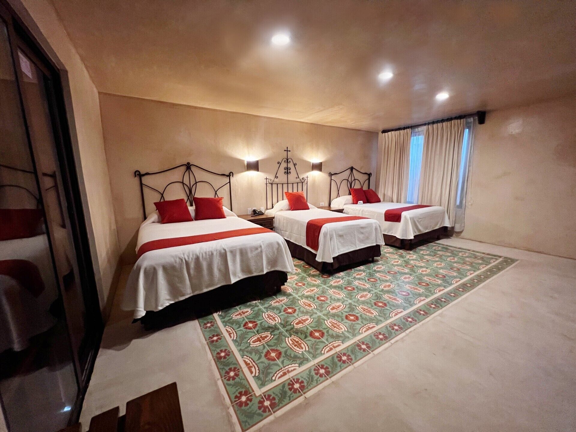 Ein Hotelzimmer mit drei Betten, jedes mit roten Kissen und weißer Bettwäsche. Im Zimmer befindet sich ein gemustertes Teppich und ein großes Fenster mit Vorhängen.