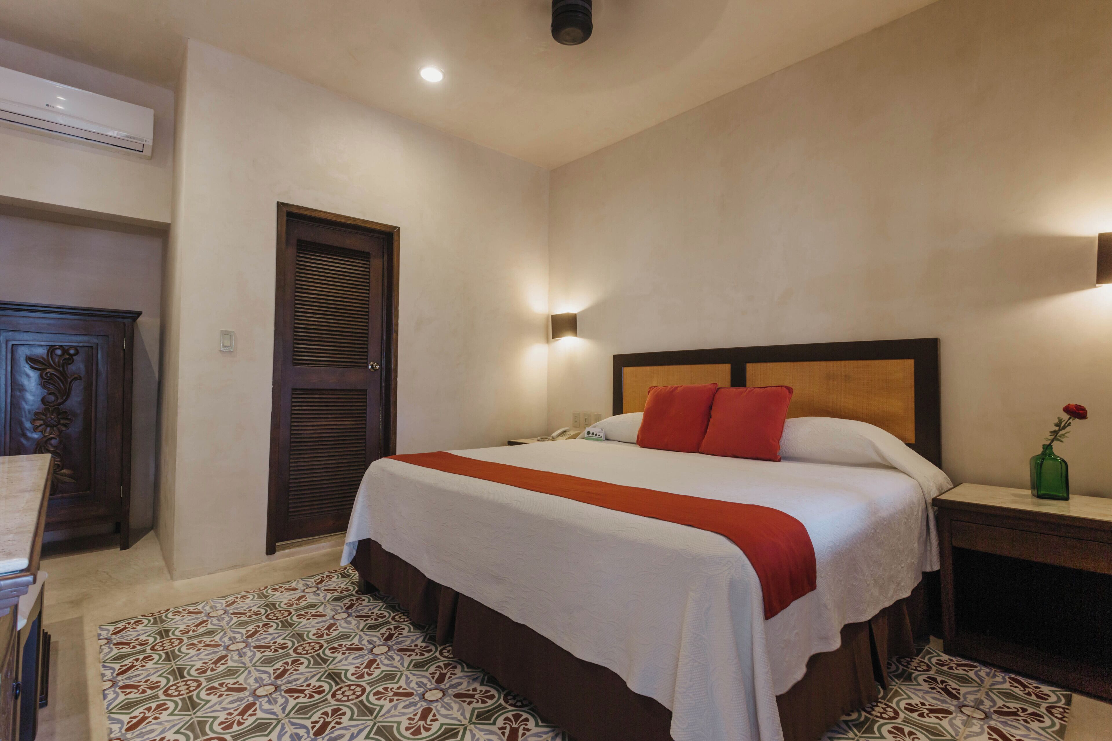 A hotel room with a bed, a nightstand, a lamp, a vase with a flower, and a patterned rug.
