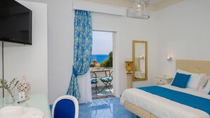 Habitación de hotel con cama, televisión, escritorio con silla, mesa con sillas, balcón con vista al mar y espejo en la pared.