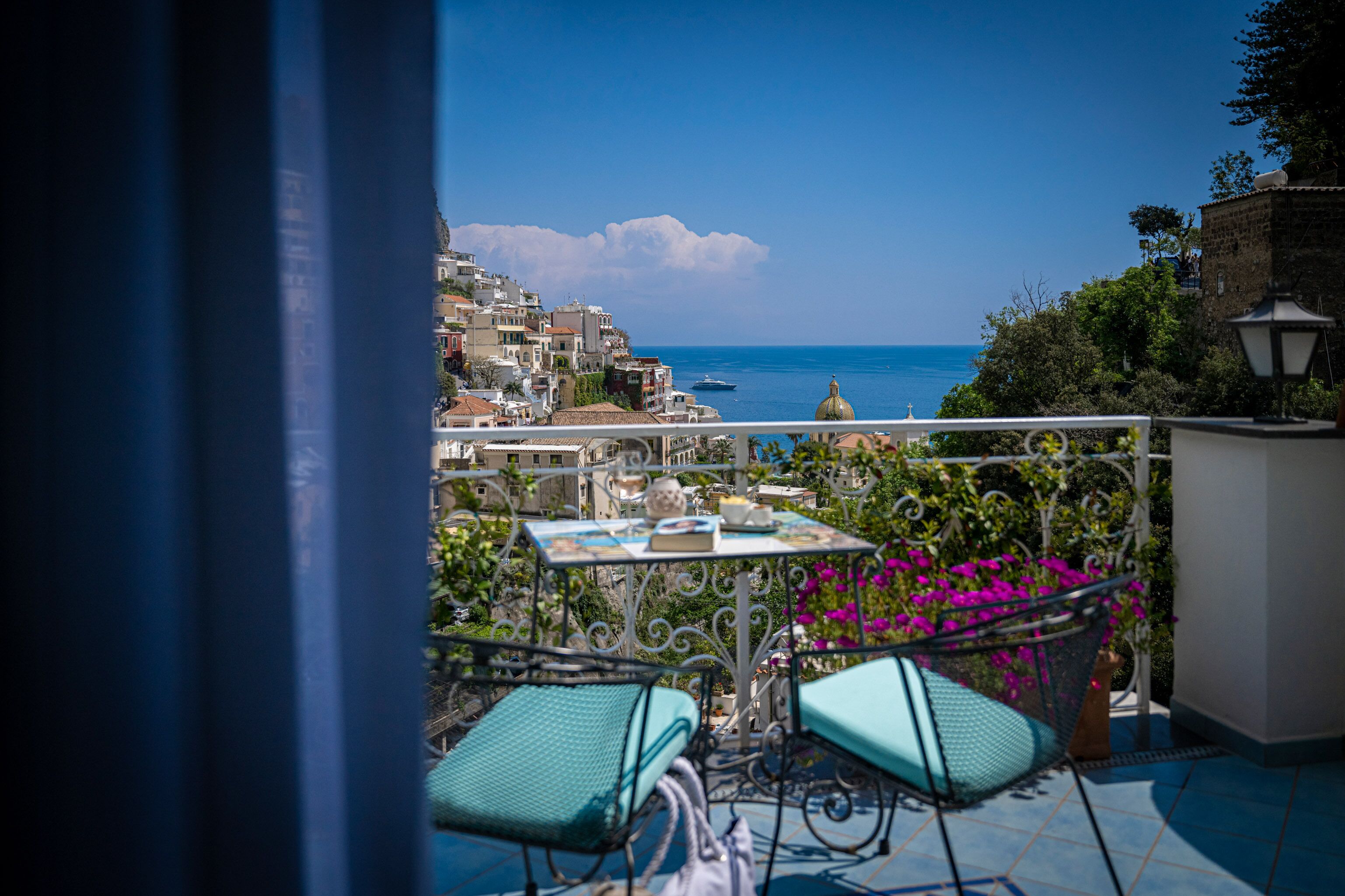 A balcony with a table and chairs, overlooking a coastal town with buildings and the sea.