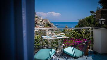 A balcony with a table and chairs, overlooking a coastal town with buildings and the sea.