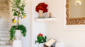 Una sala de estar con un sofá, un jarrón blanco con flores rojas, una planta en maceta y un espejo en la pared.