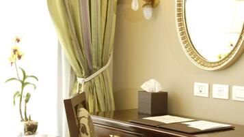 A hotel room with a wooden desk, a chair with floral upholstery, a round table, a potted orchid, a mirror with an ornate frame, a wall-mounted lamp, and a window with green curtains.