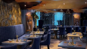 Interior de restaurante con asientos tapizados en azul oscuro, mesas dispuestas con vajilla en tonos dorados y una lámpara de araña moderna.