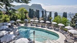 Un espace piscine doté de chaises longues et de parasols, entouré d’arbres et offrant une vue sur une étendue d’eau, avec des montagnes en arrière-plan.