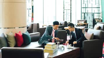 Two men in business attire sitting on a leather couch in a well-lit room with a large pillar, books, and a glass on the coffee table.