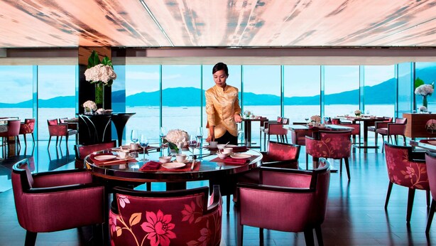 Un restaurant avec des tables rondes et des chaises rouges, un serveur en tenue traditionnelle, et une vue sur la mer par de grandes fenêtres.