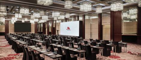 A conference room with rows of tables and chairs, a large screen, and chandeliers.