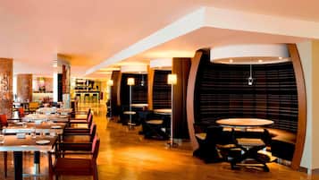 A modern restaurant with a long counter and multiple tables, featuring a warm, inviting atmosphere with wooden flooring and contemporary lighting fixtures.