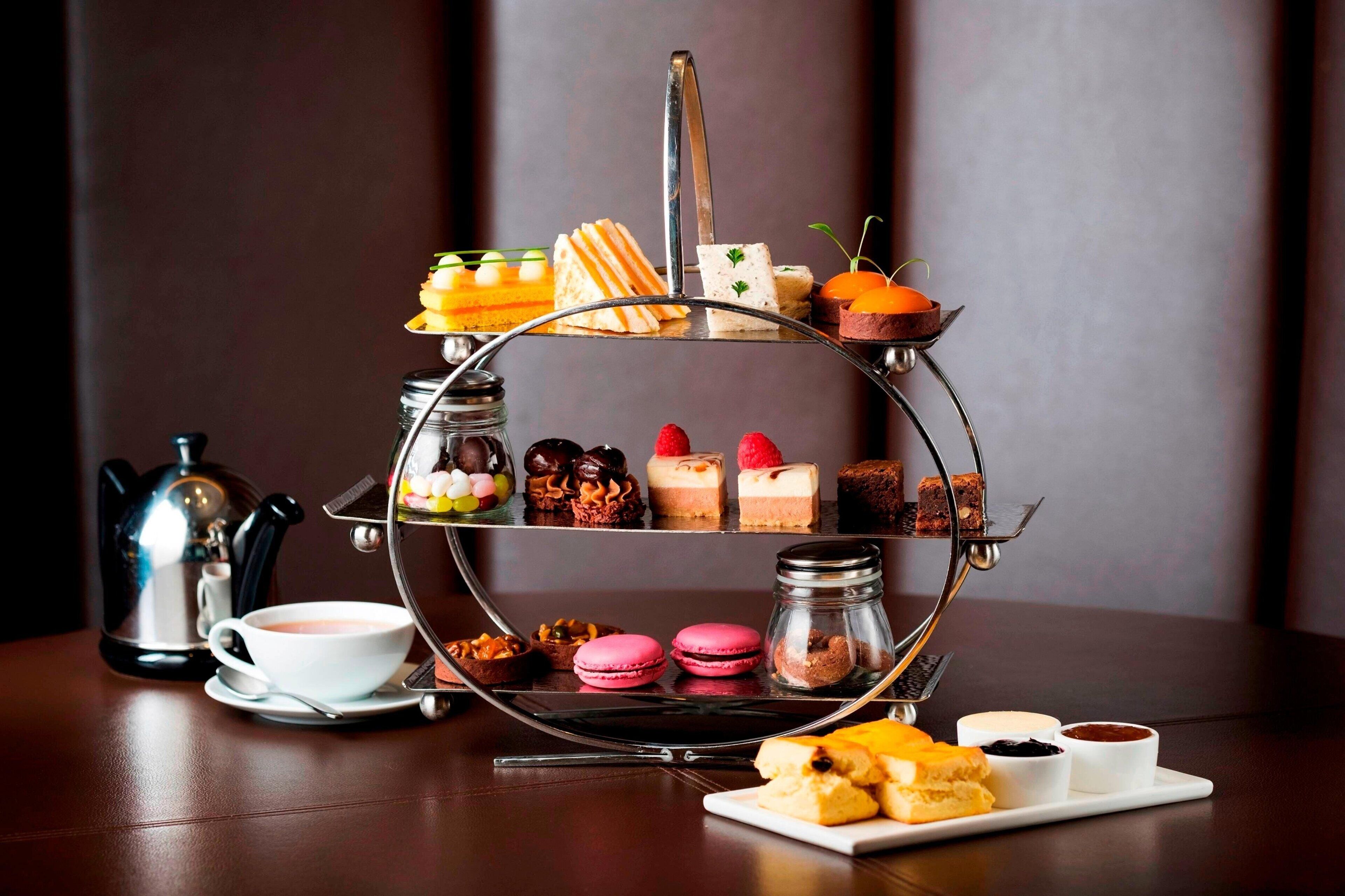 A three-tiered dessert stand with various pastries and desserts, a coffee cup, and a teapot on a table.