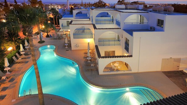 A swimming pool adjacent to a building with multiple balconies and a view of the sea. - Petrosana Hotel Apartments (Ayia Napa)