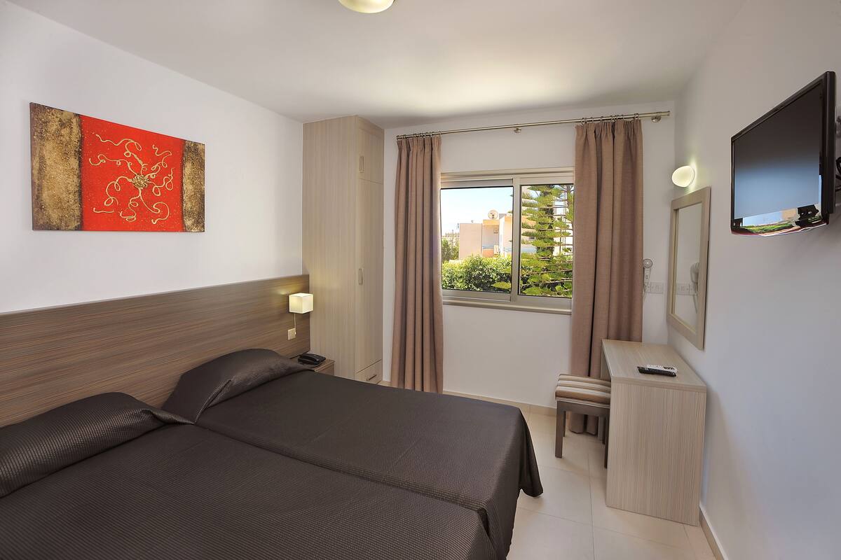 Hotellihuone, jossa on kaksi sänkyä, työpöytä, tuoli, televisio, ikkuna verhoilla ja taulu seinällä.