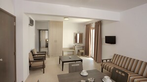 A modern living room with a sofa, a dining table with chairs, a TV mounted on the wall, and a kitchen area in the background. - Petrosana Hotel Apartments (Ayia Napa)