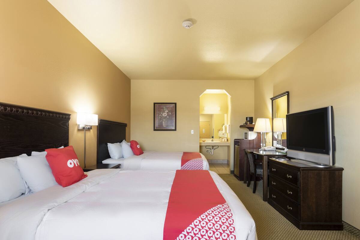 Habitación de hotel con dos camas, un escritorio, televisión y un baño visible a través de un arco.