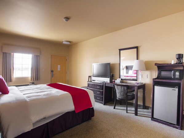 Une chambre d’hôtel équipée d’un lit, d’un bureau avec un ordinateur, d’une télévision, d’un miroir, d’une machine à café et d’un petit réfrigérateur.