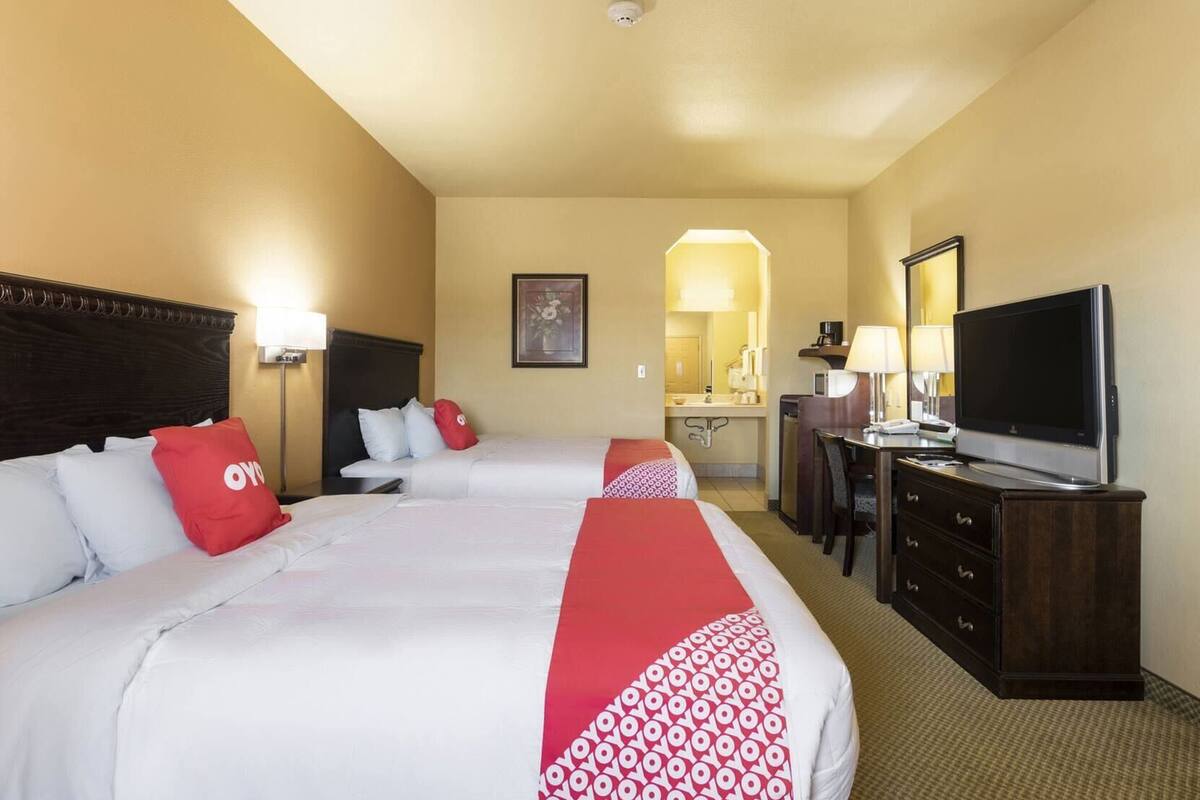 Habitación de hotel con dos camas, un escritorio con televisor, un espejo y un baño visible a través de un arco.