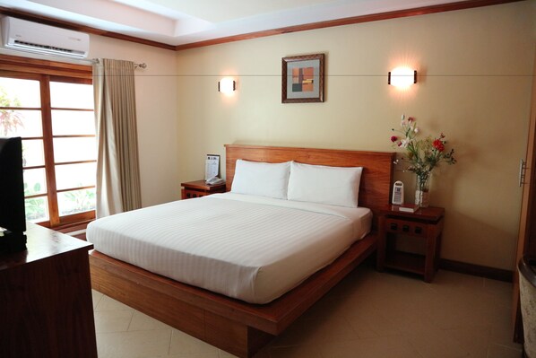 Una cama bien hecha en una habitación de hotel, con cabecera de madera, una mesita de noche con lámpara y una pequeña mesita con un florero.