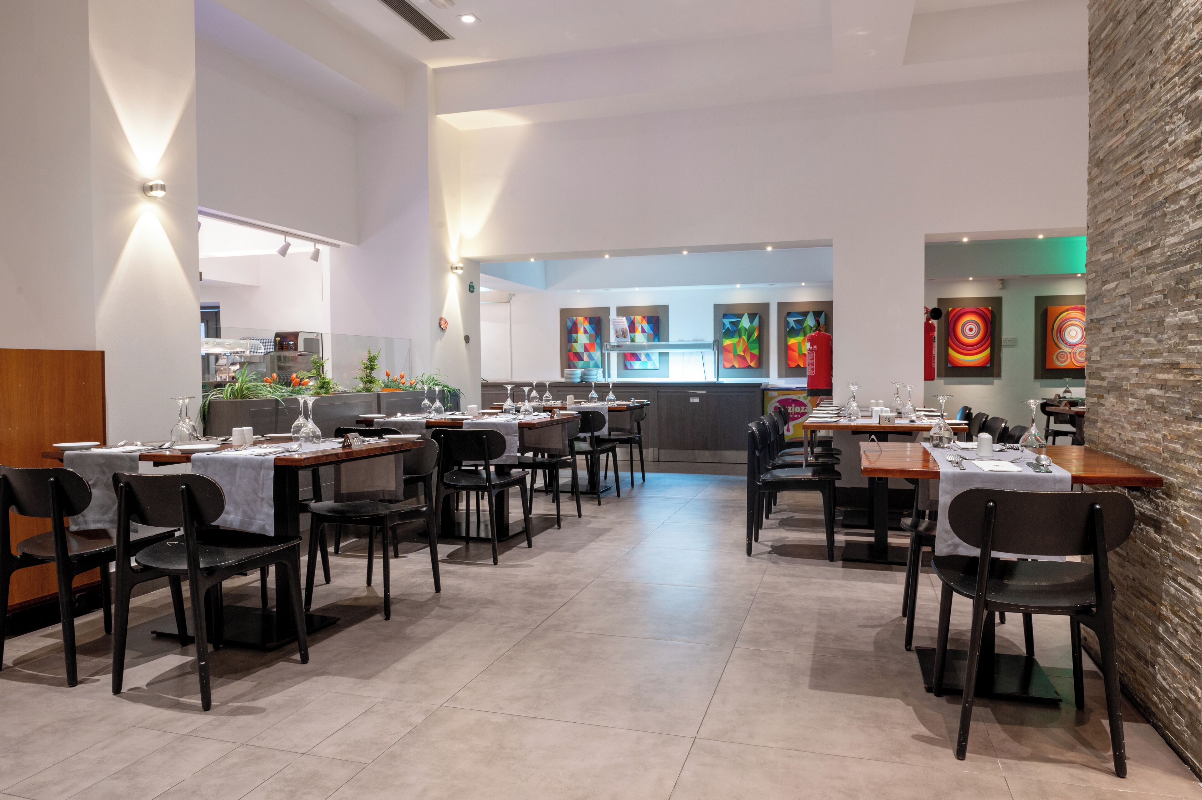 A modern restaurant interior with multiple dining tables and chairs, a kitchen area in the background, and colorful artwork on the walls.
