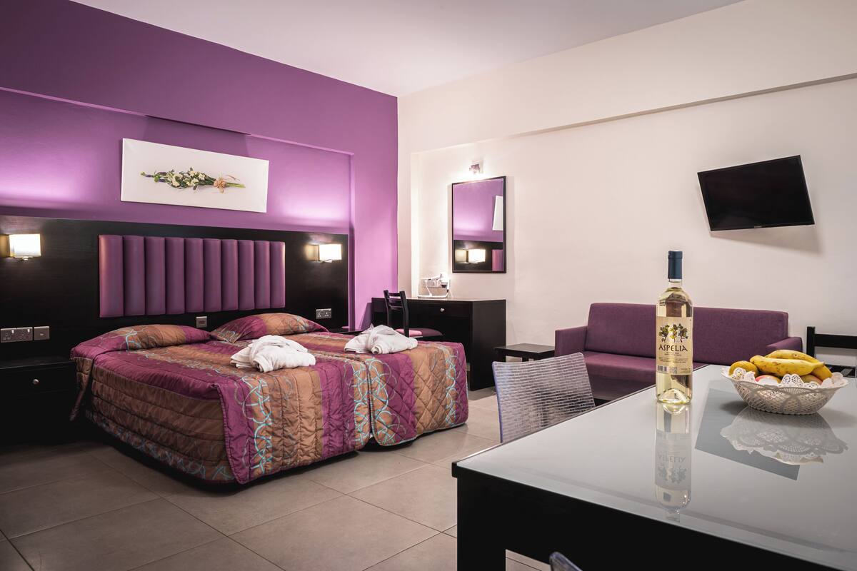A modern hotel room with a large bed, a sofa, a dining table, and a TV.