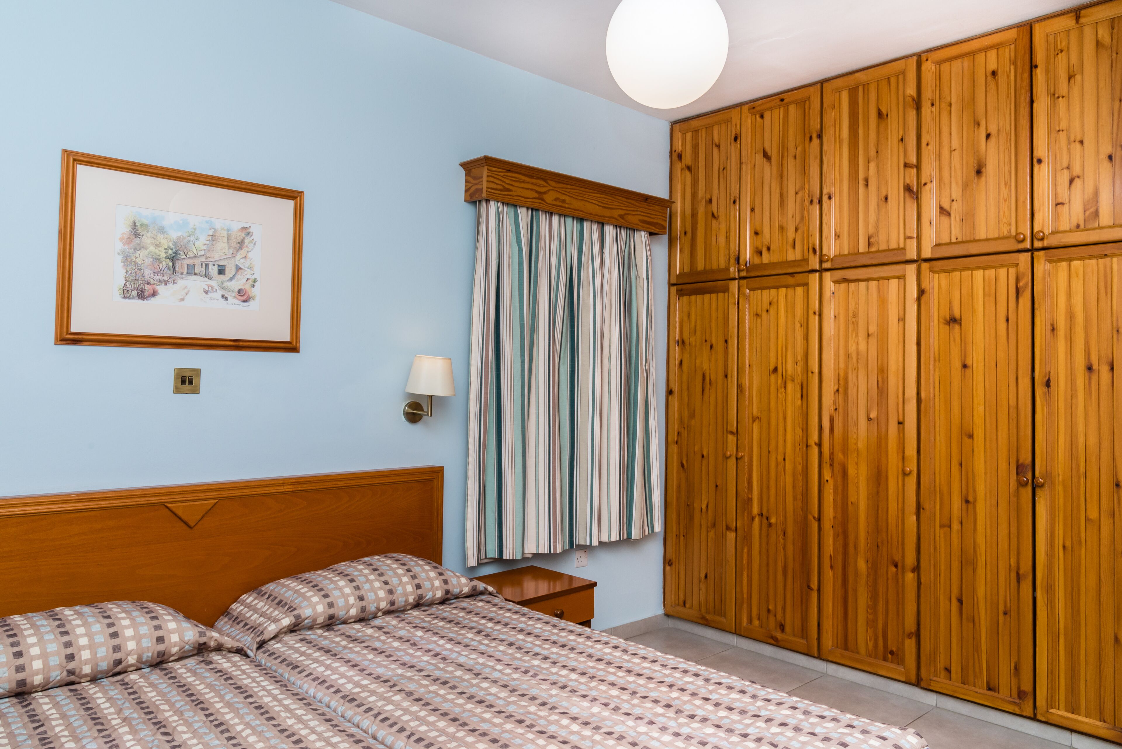 Un dormitorio con una cama de madera, una mesita de noche, una lámpara de pared, un cuadro enmarcado y un armario de madera grande.