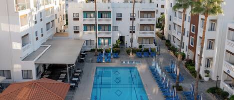 A hotel with a swimming pool surrounded by lounge chairs and umbrellas, with palm trees and a view of the city.