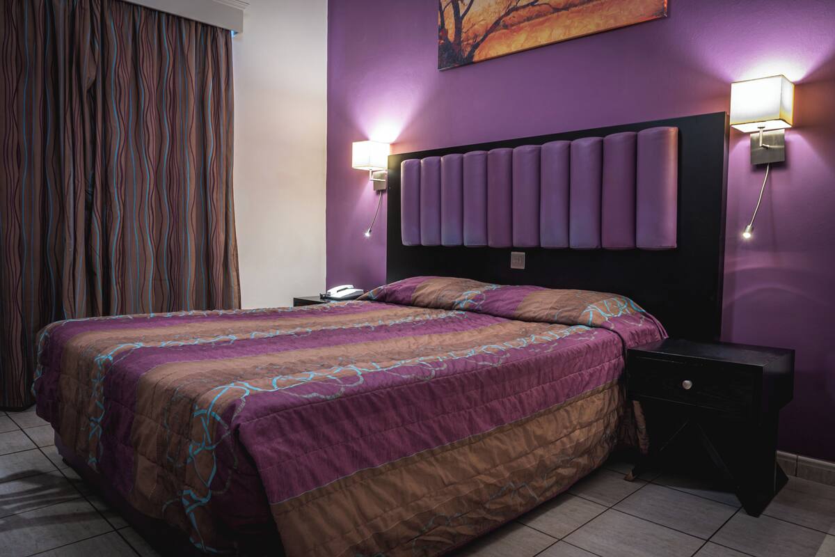 A hotel room with a large bed, a headboard with lights, a nightstand, and a curtain.