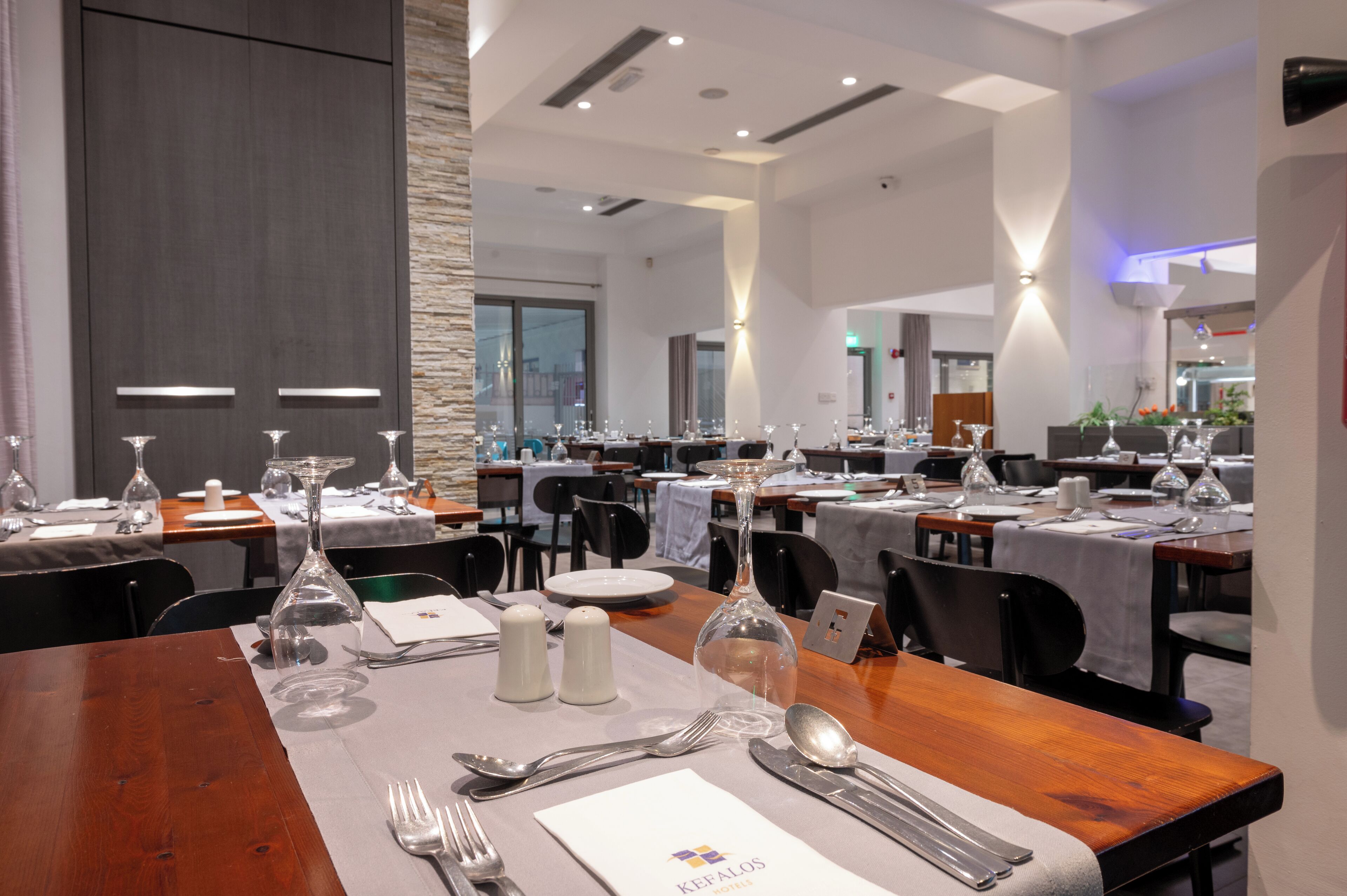 Une salle à manger avec plusieurs tables dressées pour le service, dotée de tables en bois poli, de nappes blanches et de couverts disposés avec soin.
