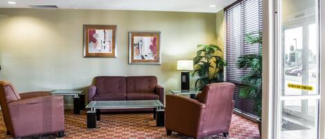 A lobby with a sofa, armchairs, a coffee table, framed artwork, a lamp, and a large window with blinds.