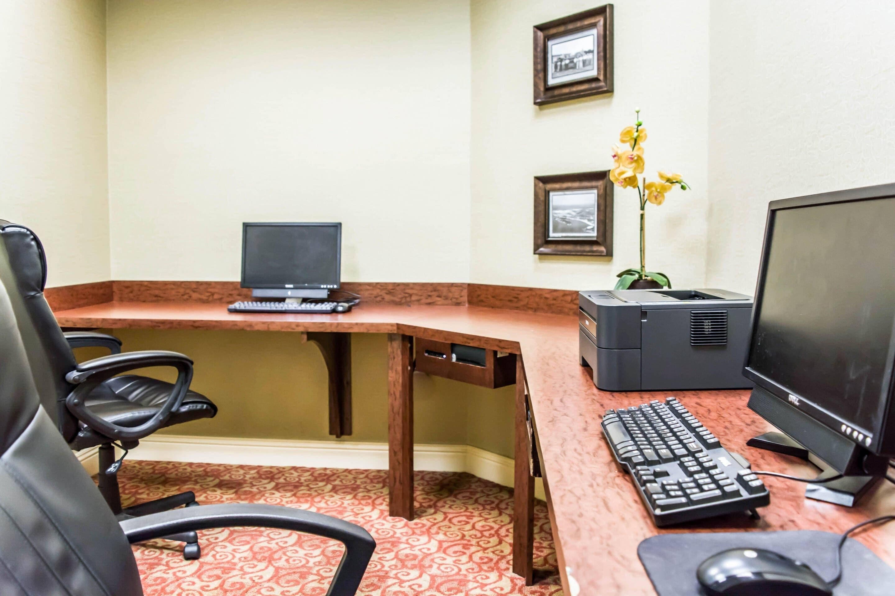 Oficina de hotel con escritorio, dos computadoras, teclado, ratón y una silla.