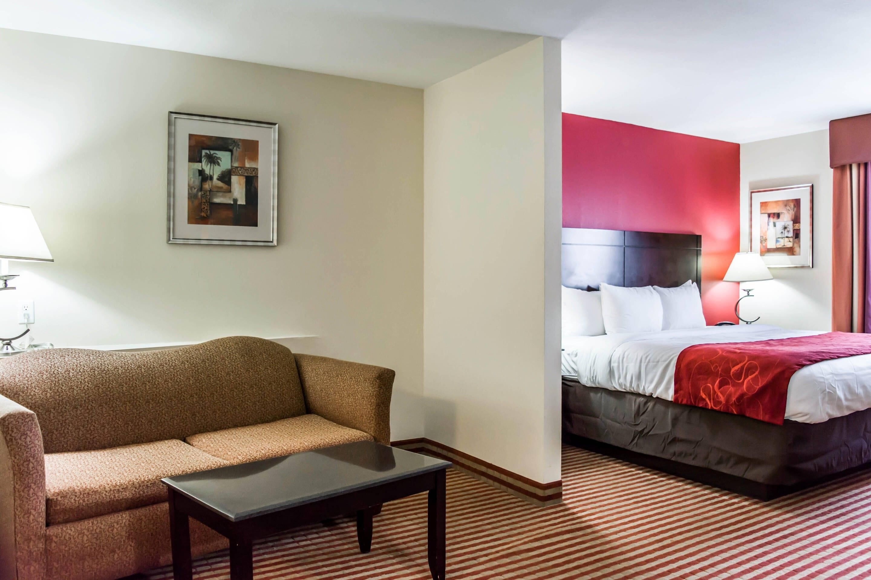 A hotel room with a sofa, a small table, a bed with a red headboard, and a framed picture on the wall.