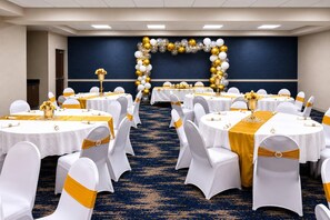 Une salle de conférence avec des tables rondes, des chaises blanches ornées de rubans dorés et une arche décorative faite de ballons.