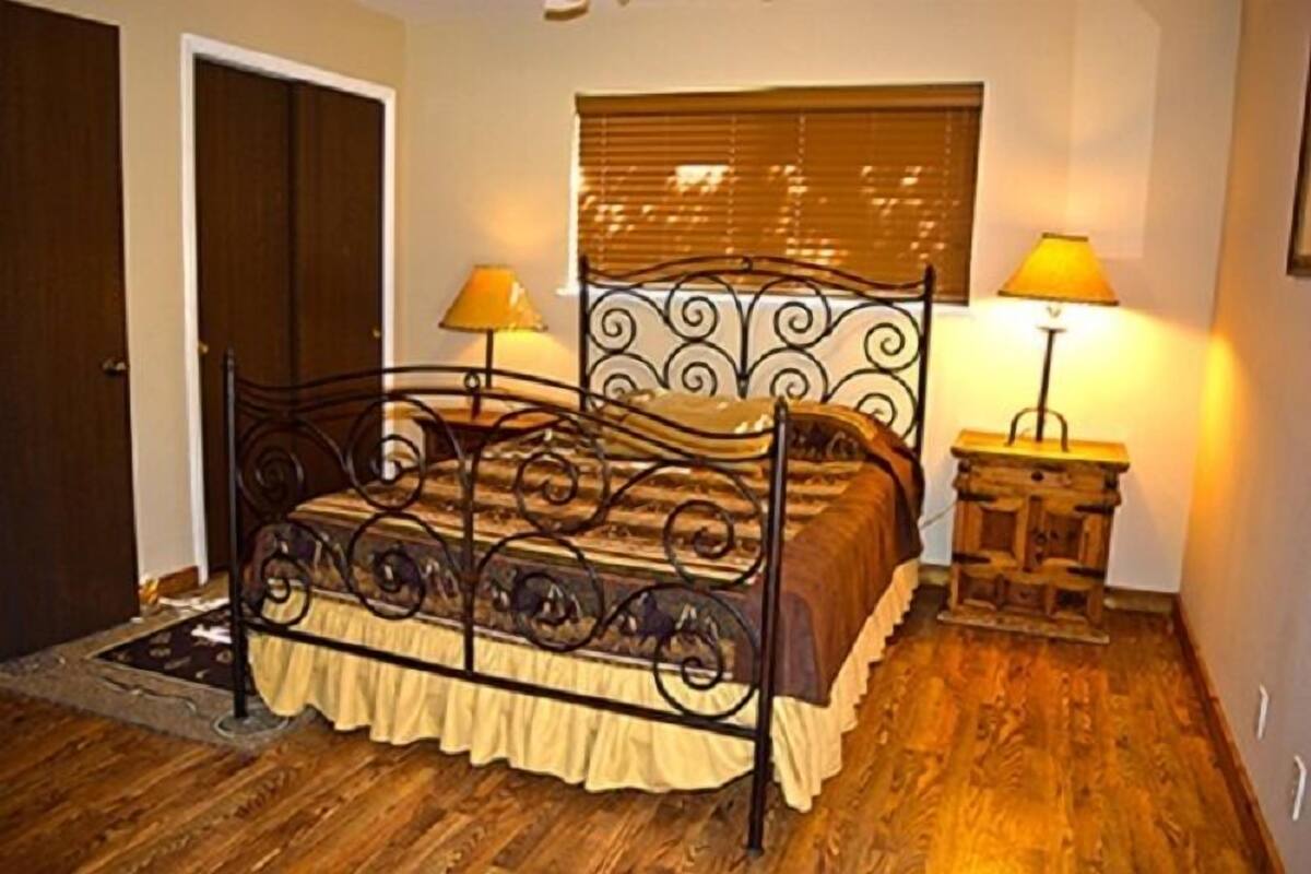 Un dormitorio con estructura de cama metálica, dos mesitas de noche con lámparas, suelo de madera y ventana con persianas.