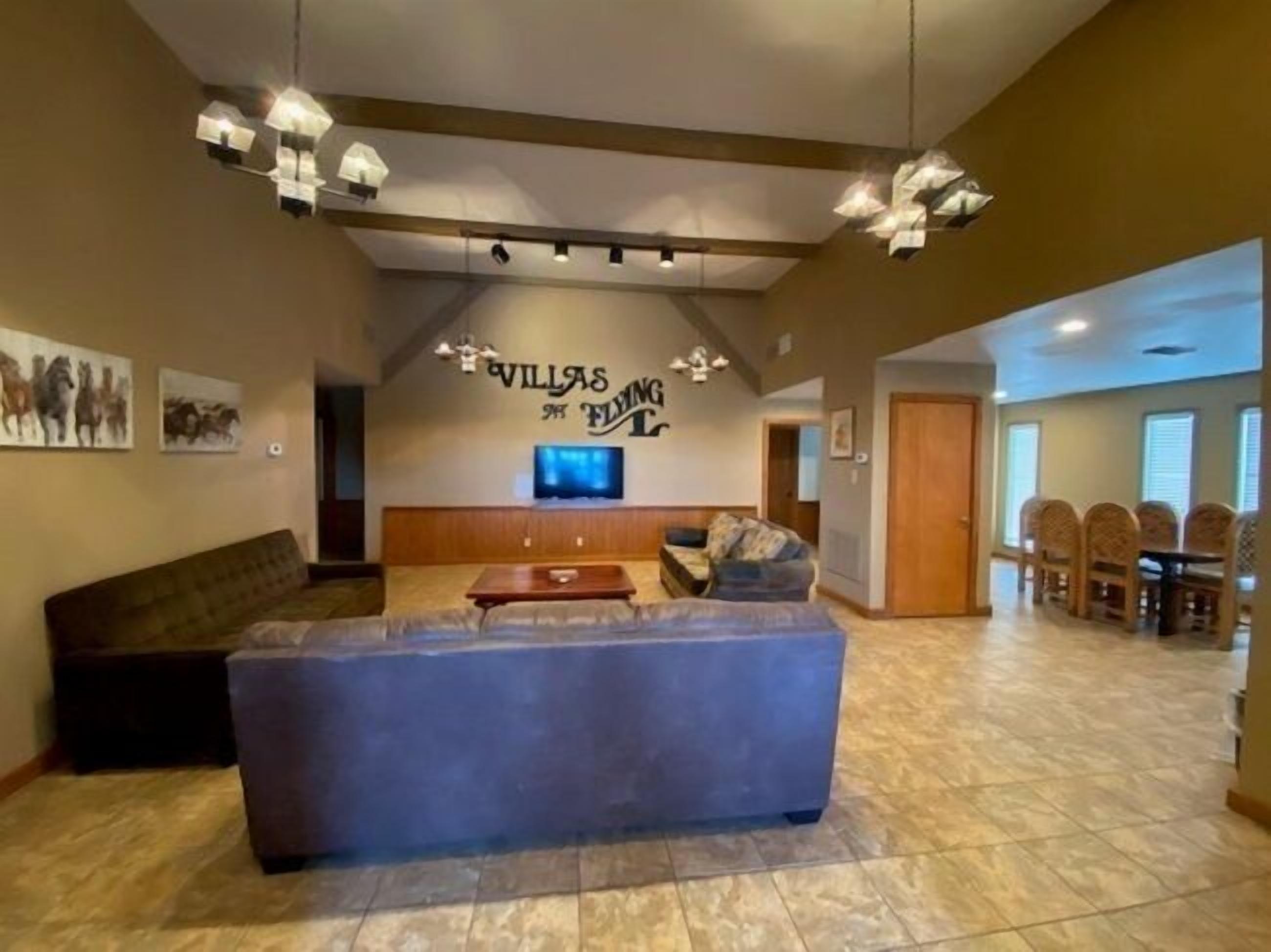 A spacious living area with a large sofa, a flat-screen TV mounted on the wall, and a dining area with wooden chairs and a table.