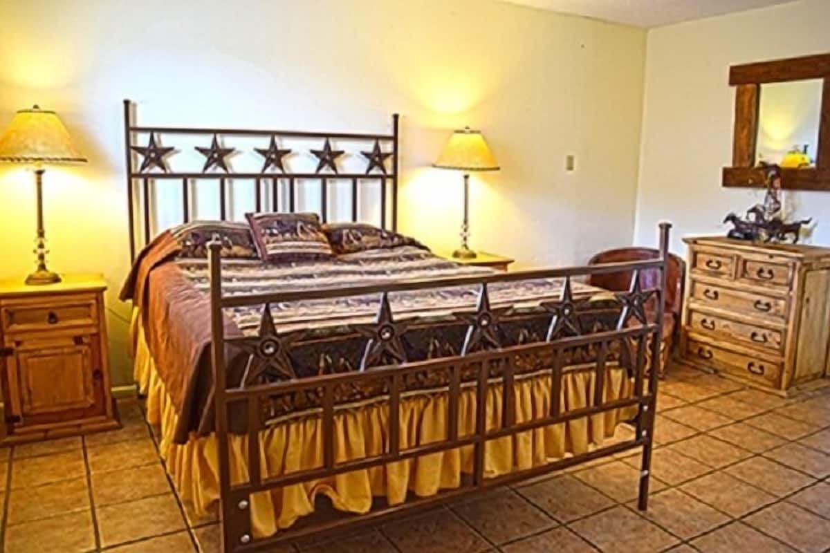 Un dormitorio con estructura de cama metálica, dos mesitas de noche con lámparas, un aparador de madera y un espejo en la pared.